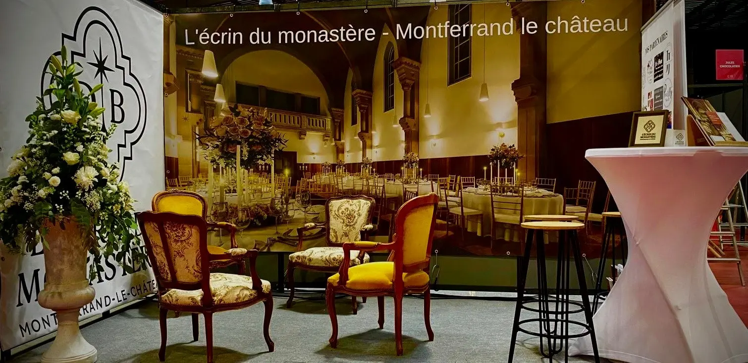 Stand de L’Écrin du Monastère au Salon des Mariés de Besançon, avec fauteuils anciens, table haute et grand visuel de la salle de réception.