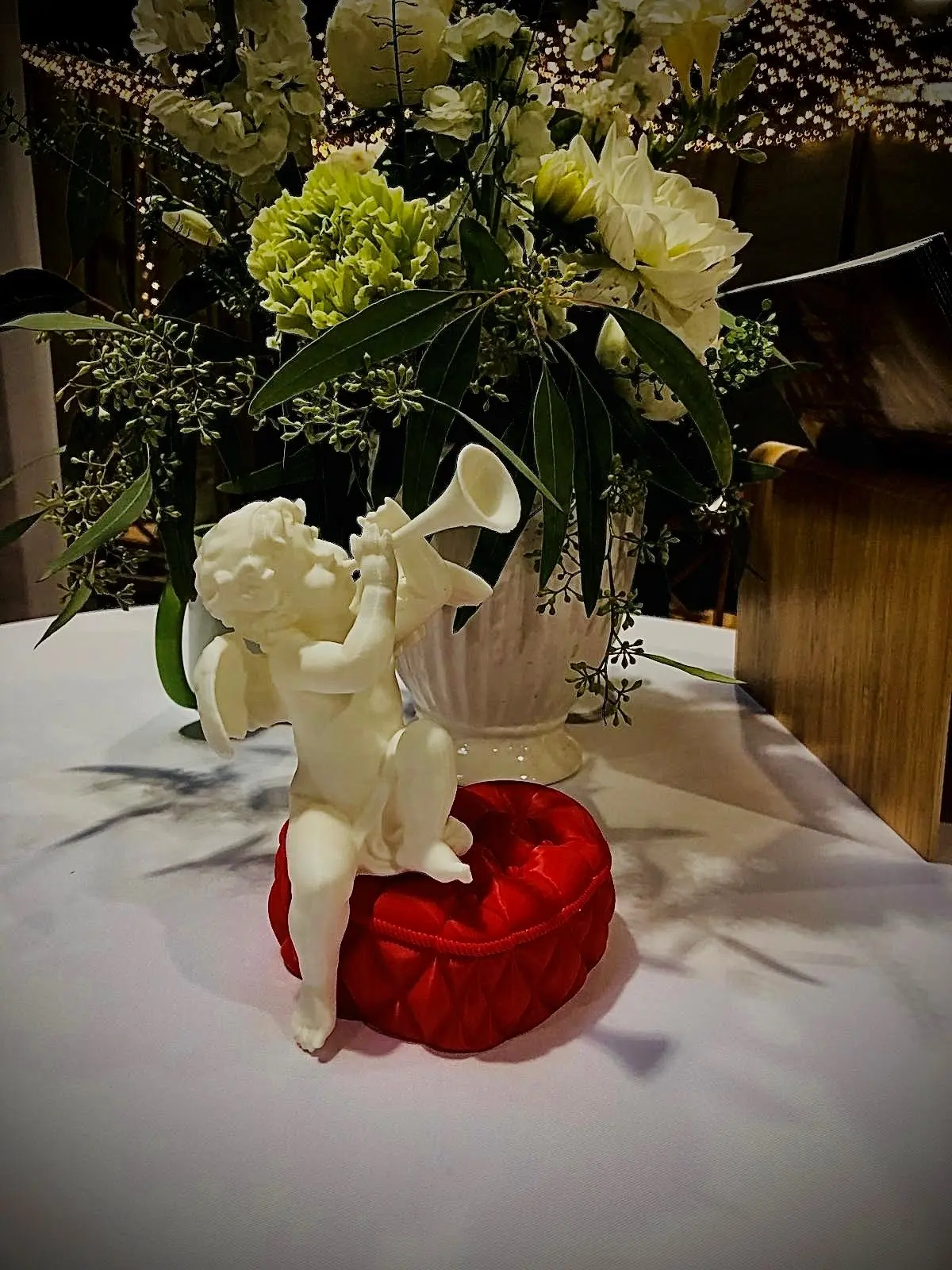 Petit angelot décoratif blanc jouant de la trompette, posé sur un coussin rouge, en centre de table fleuri sur le stand de L’Écrin du Monastère.
